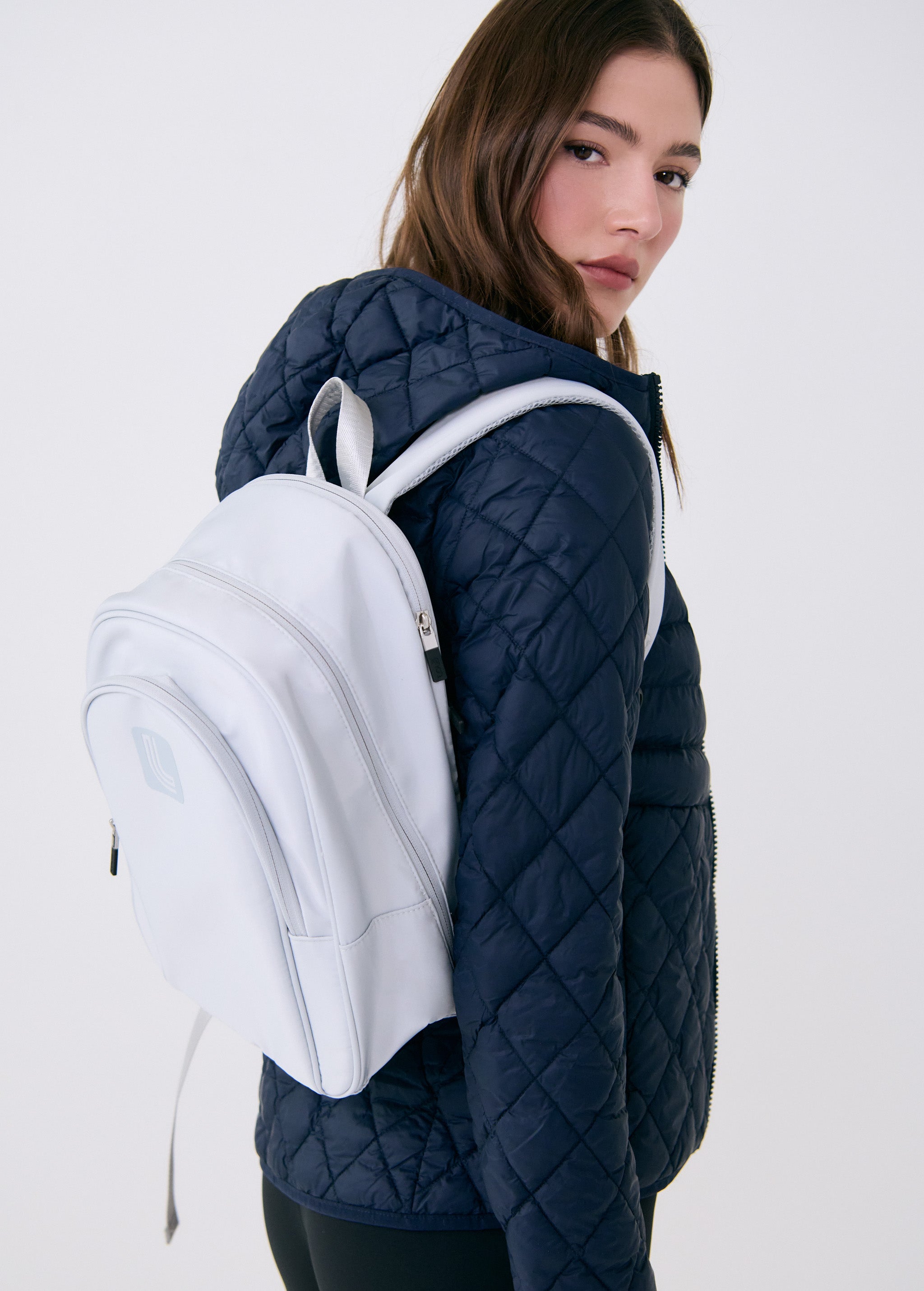 Lolë Chelsea backpack in pale grey, slung over the shoulder of a model looking back, front pocket and logo in view