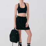 Lolë Chelsea backpack in black, held in hand beside a model in a black crop top and mini skirt with trainers