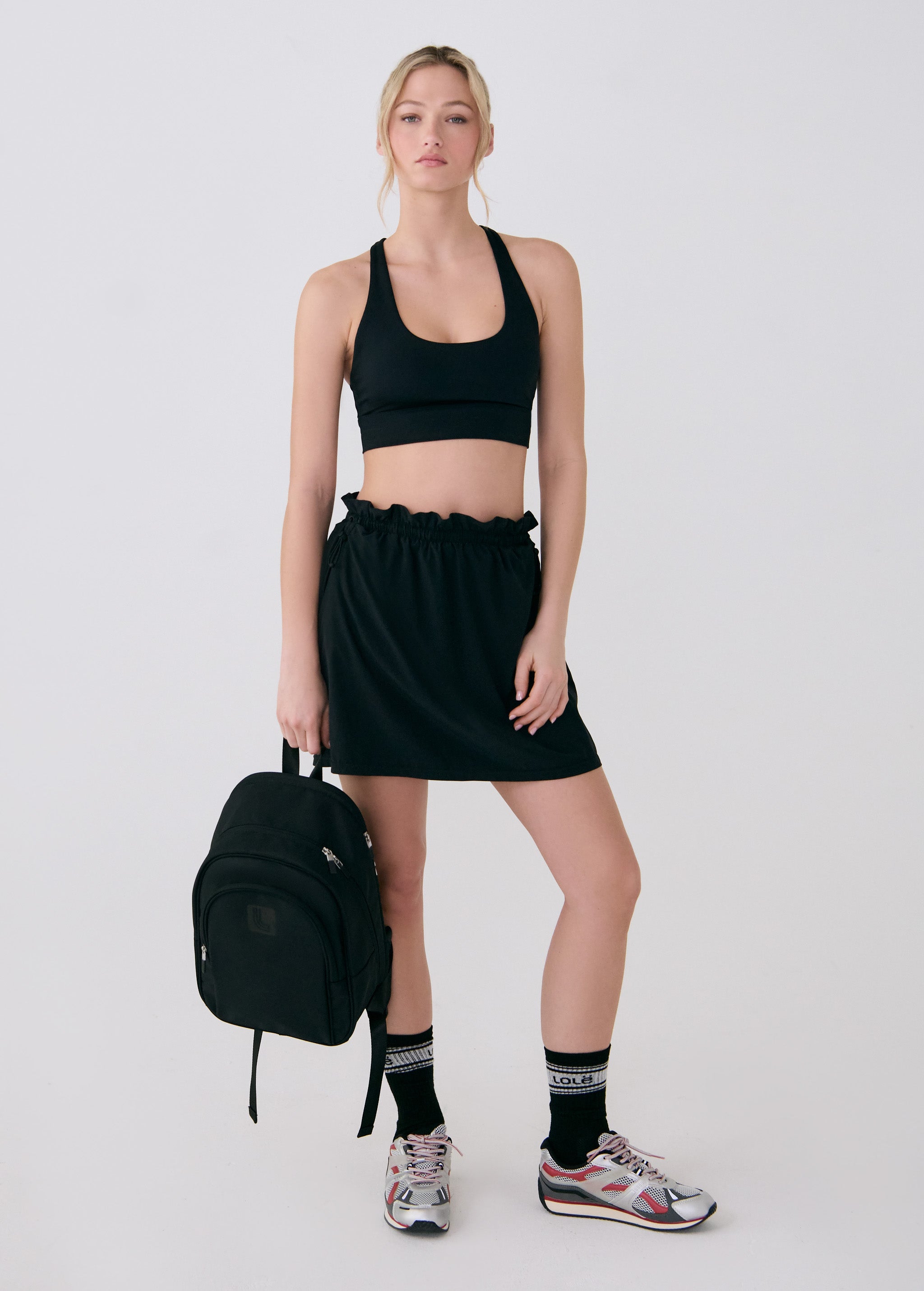 Lolë Chelsea backpack in black, held in hand beside a model in a black crop top and mini skirt with trainers