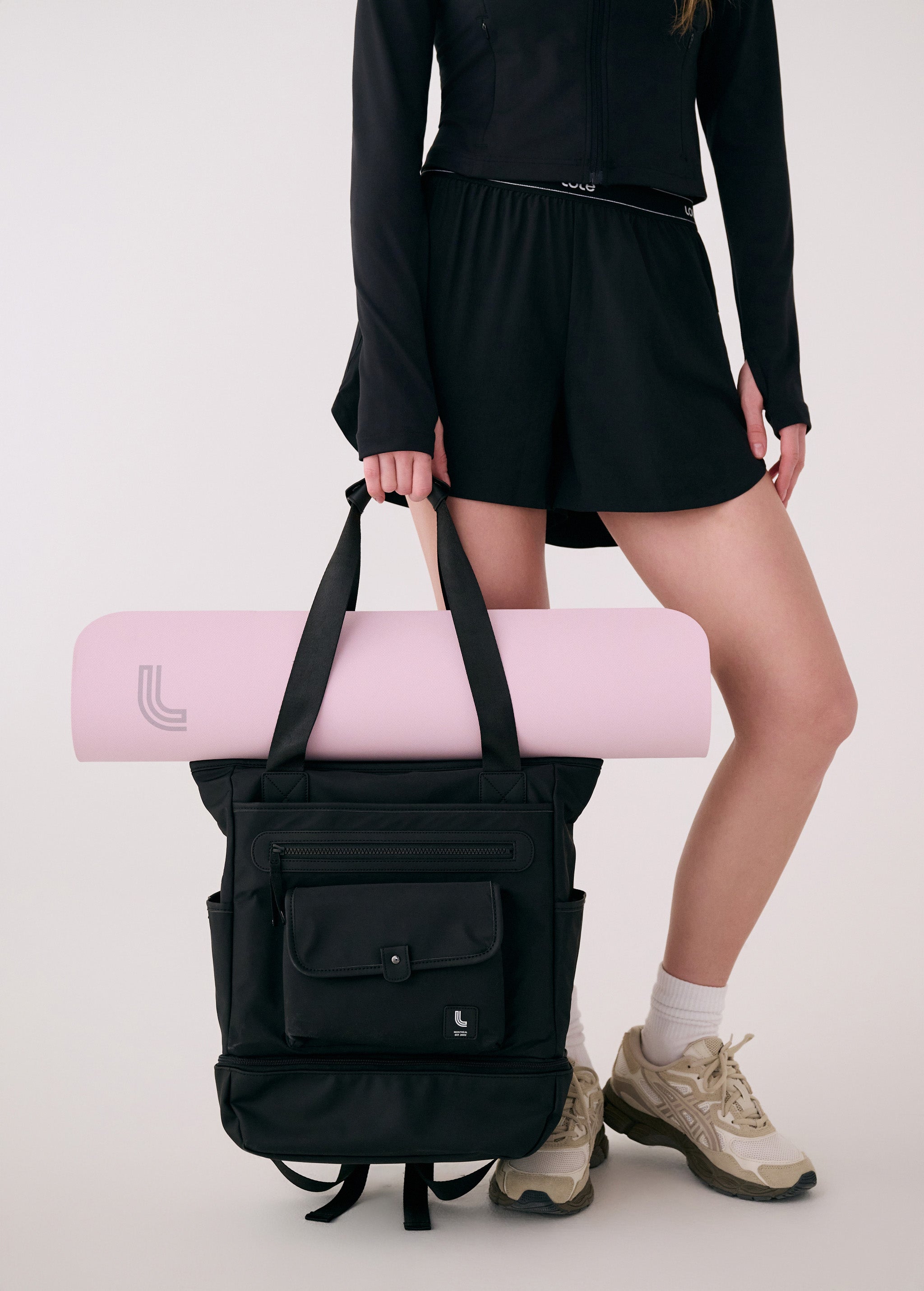 Lolë Lily 2.0 bag in black, held by a model with a pink yoga mat tucked under the tote handles, front view