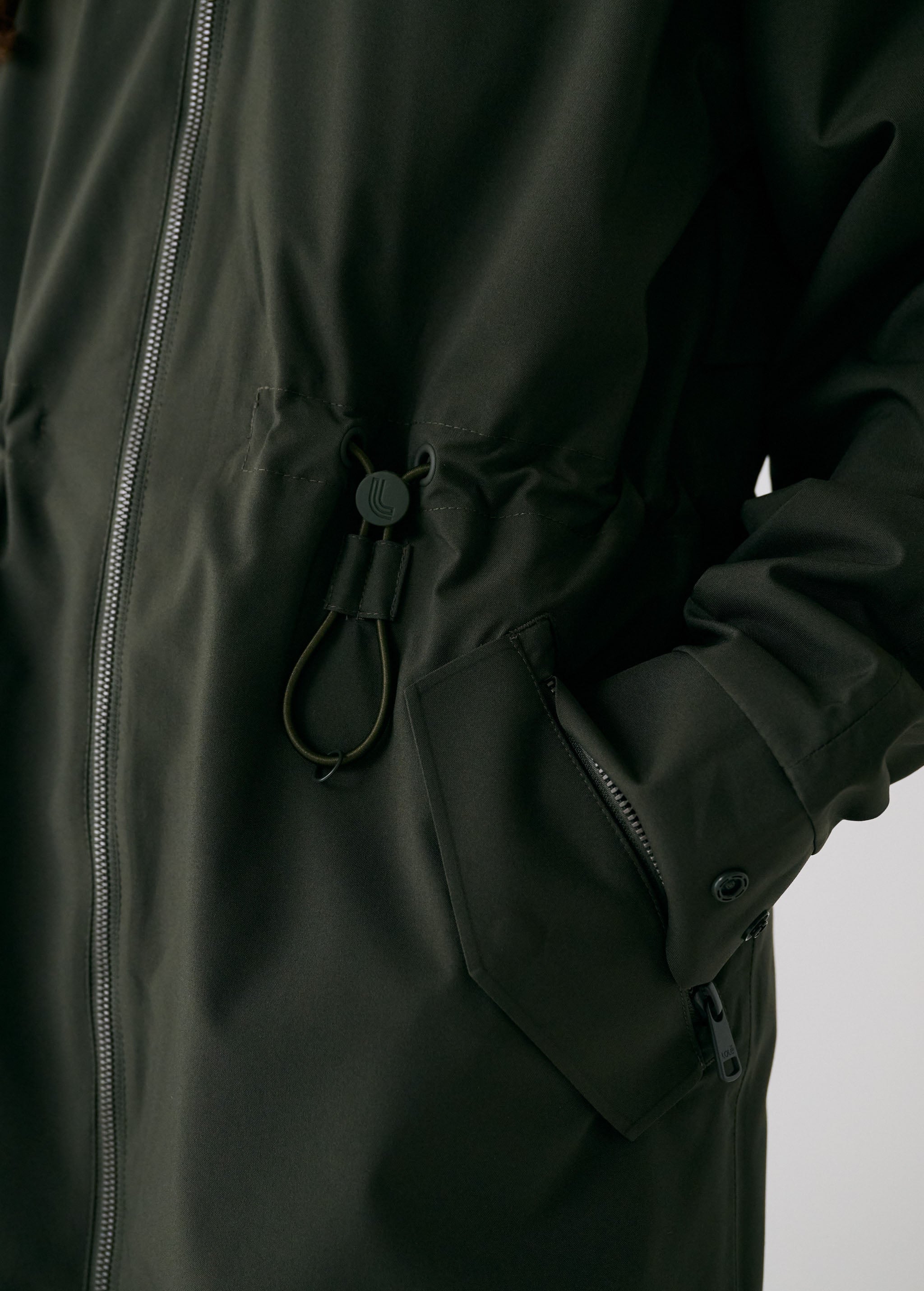 Lolë Piper 2.0 rain jacket in olive green, close view of the waist drawcord toggle and angled zip pocket