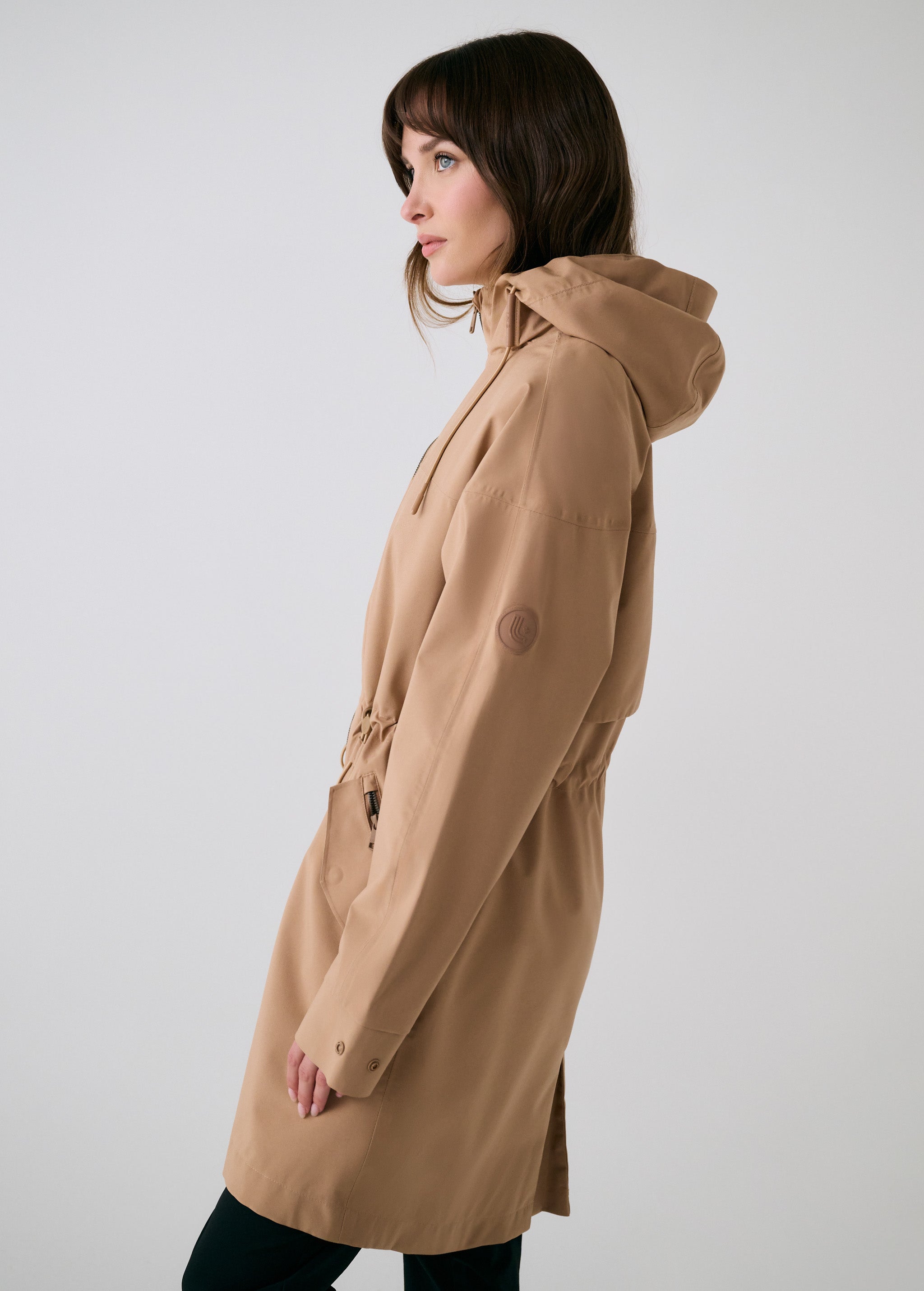 Lolë Piper 2.0 rain jacket in camel, side view showing the hood, sleeve logo and drawcord waist