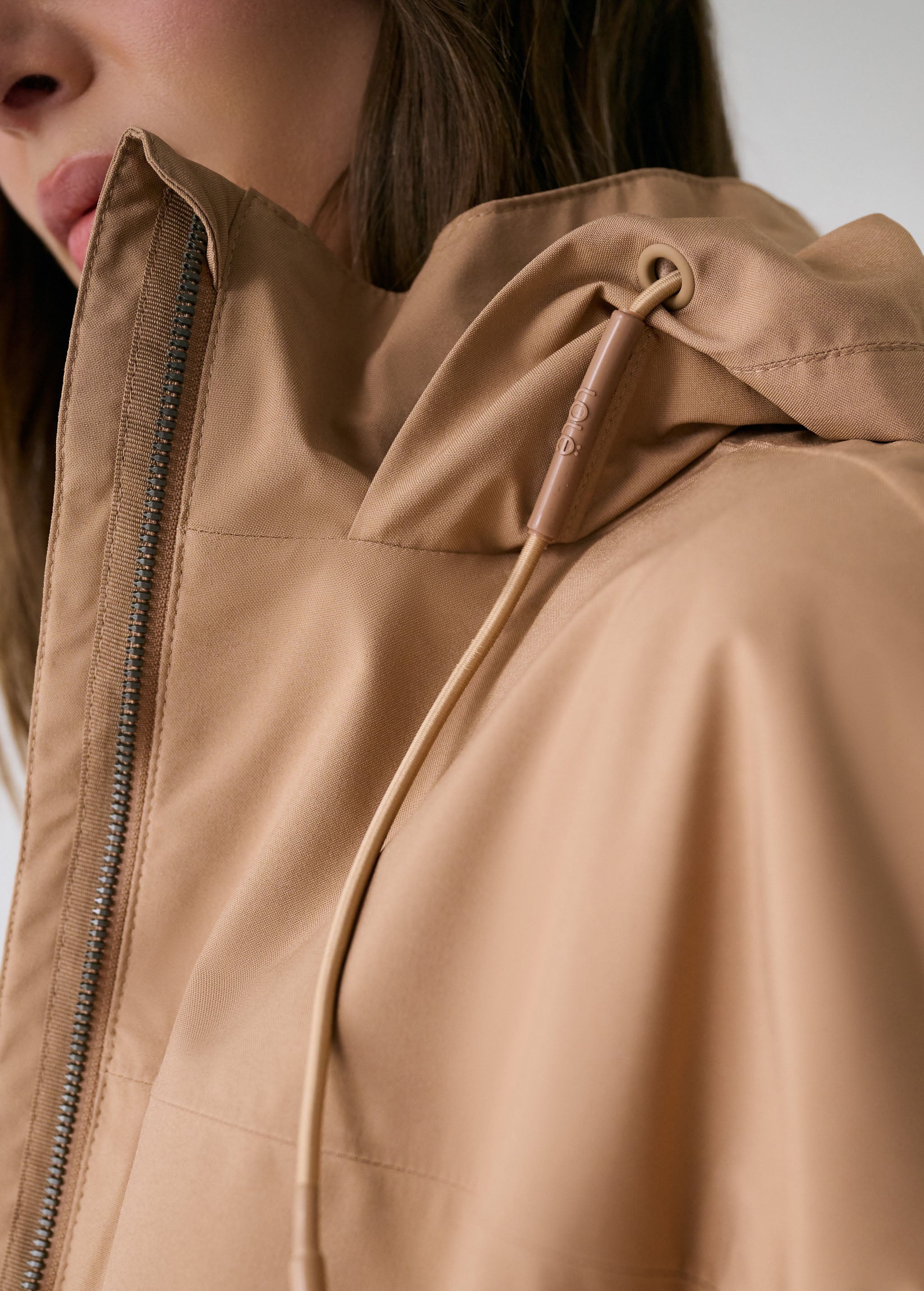 Lolë Piper 2.0 rain jacket in camel, close-up of the collar, front zip and branded hood drawcord toggle
