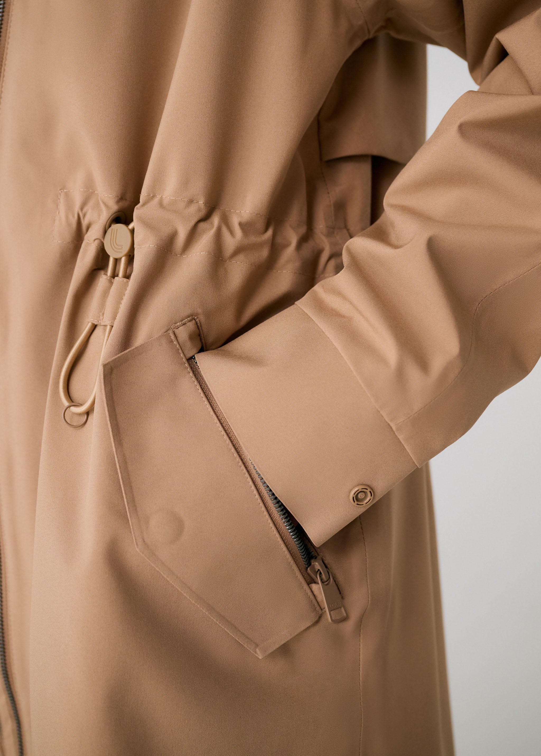 Lolë Piper 2.0 rain jacket in camel, close view of the drawcord waist toggle, flap pocket and cuff snap