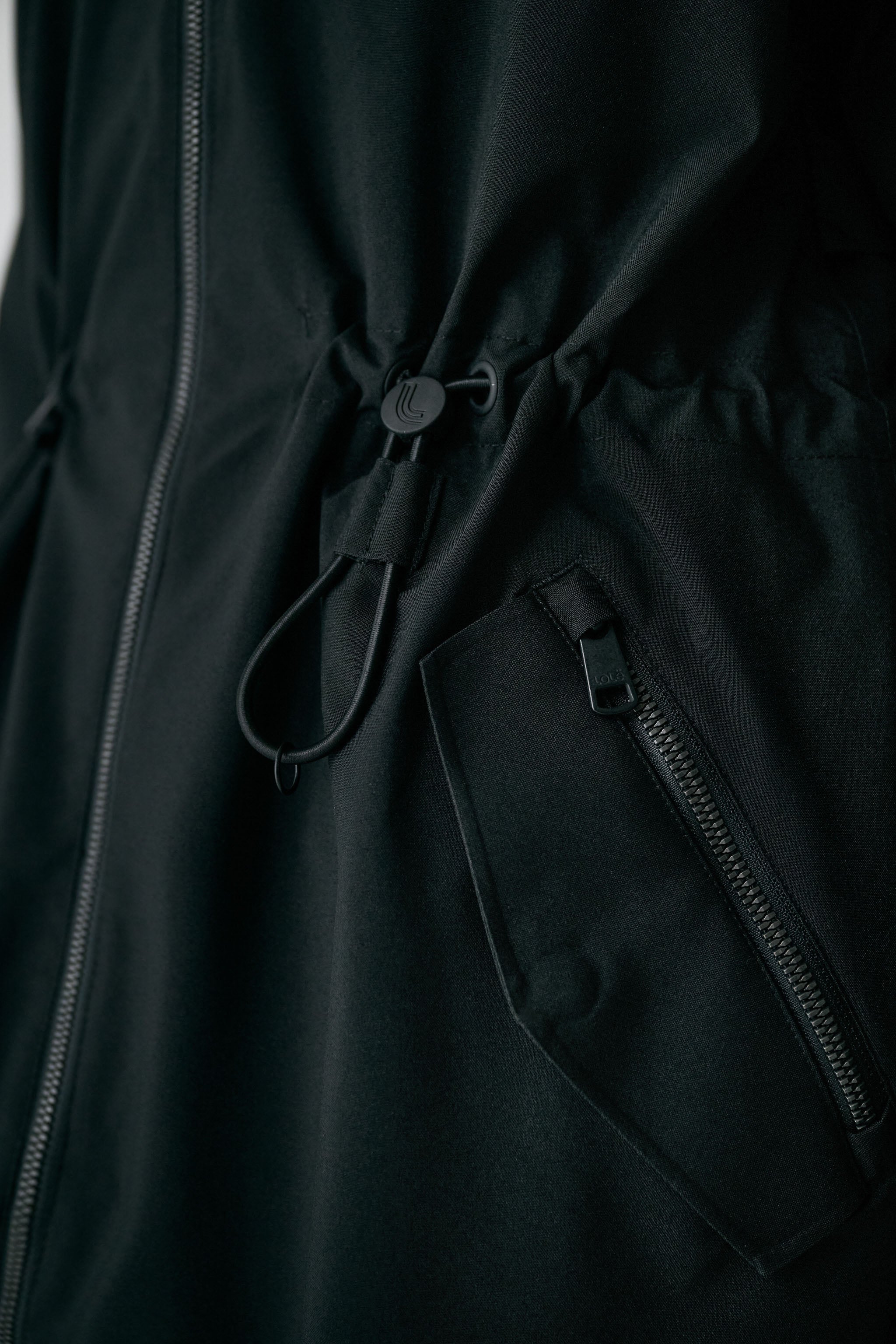 Lolë Piper 2.0 rain jacket in black, close-up of the branded drawcord waist toggle and zip hip pocket