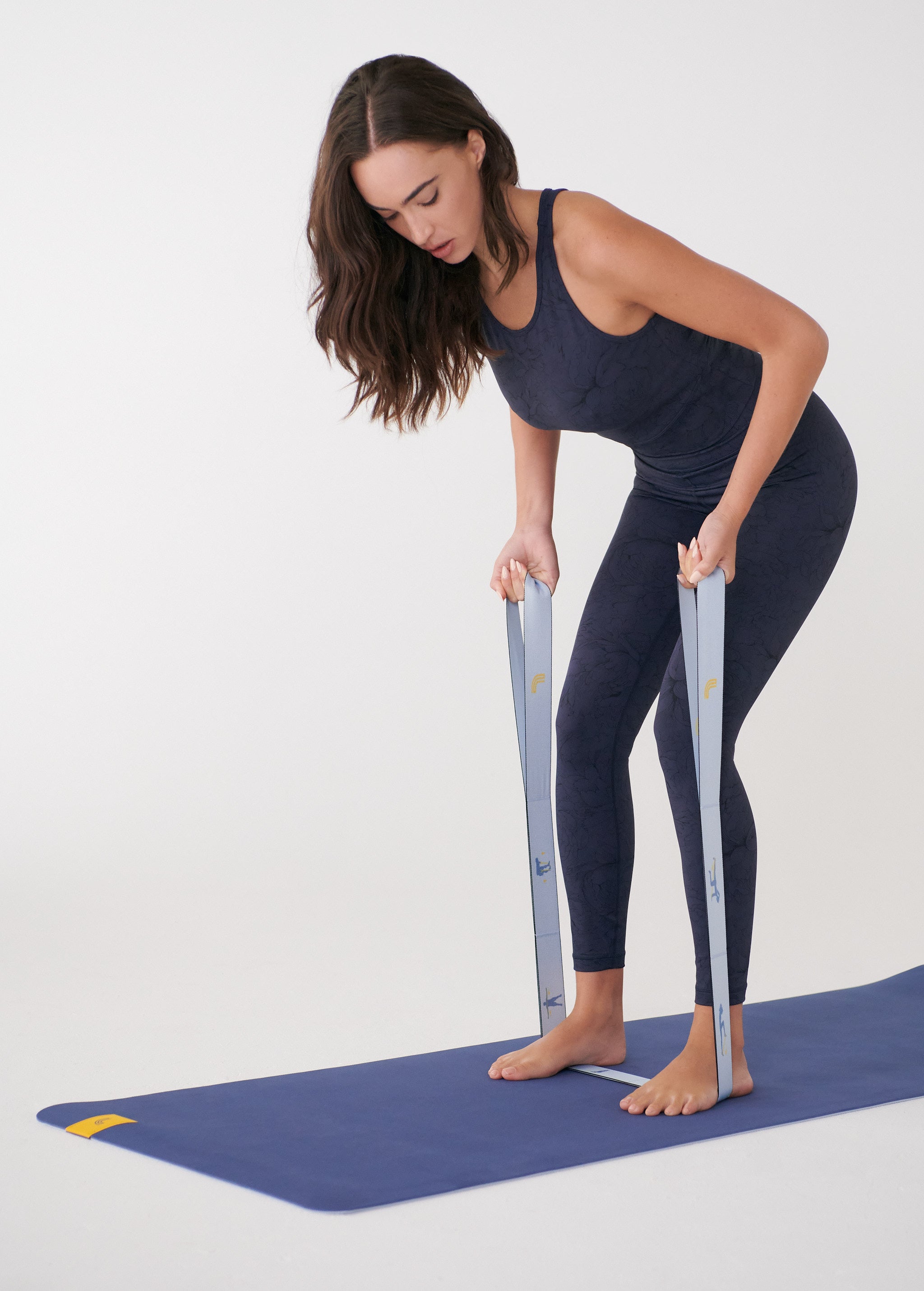 Lolë Pose yoga mat in blue, laid flat as a woman stands barefoot on it holding the pale blue strap taut