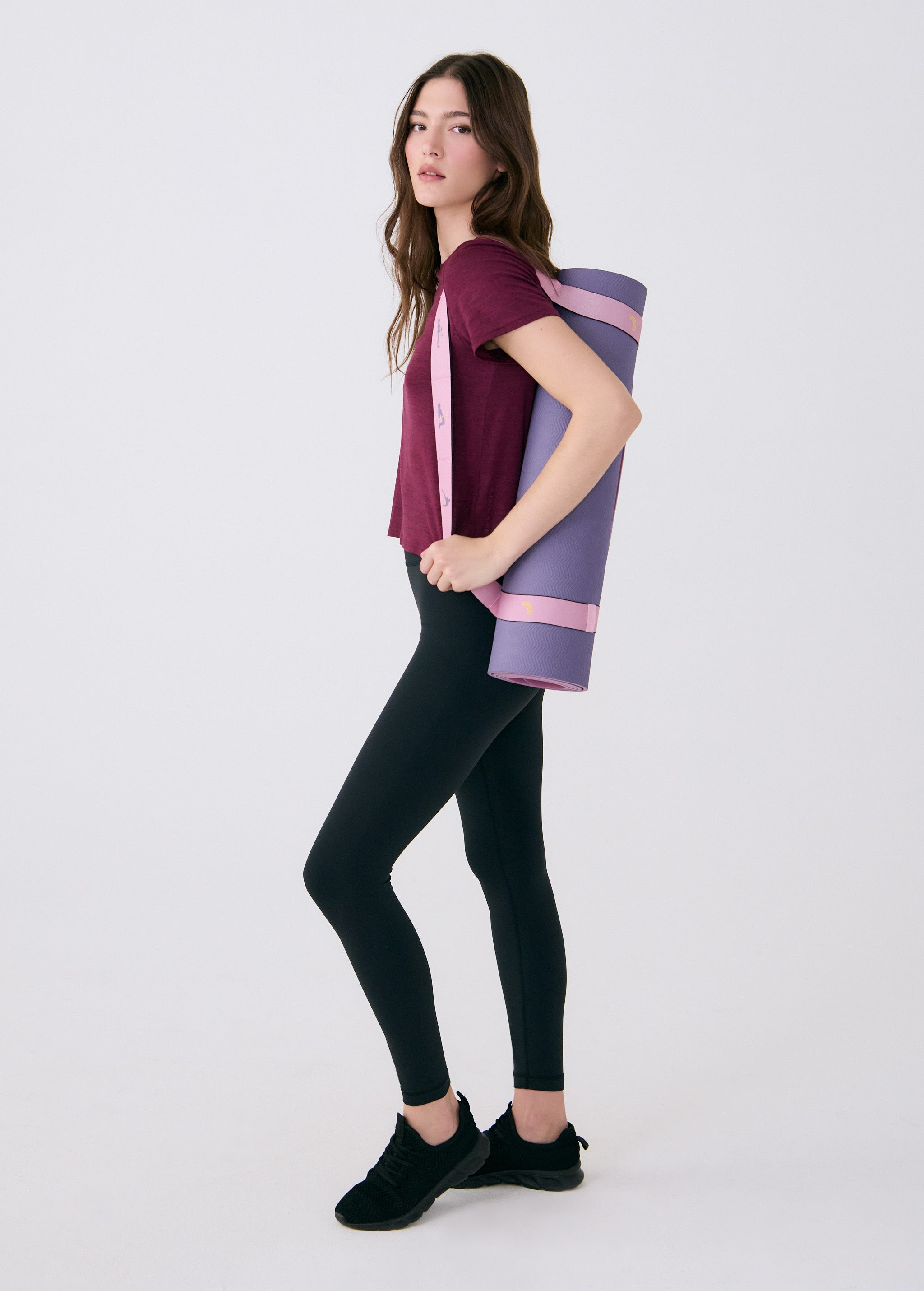 Lolë Pose yoga mat in lavender purple, a woman carries the rolled mat at her side by its pink strap