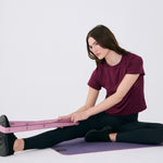 Lolë Pose yoga mat in mauve purple, a seated woman stretches with the pink strap looped around her foot