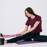 Lolë Pose yoga mat in mauve purple, a seated woman stretches with the pink strap looped around her foot