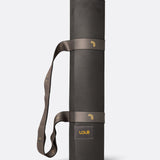 Lolë Pose yoga mat in charcoal, rolled up with the taupe shoulder strap looped for carrying