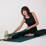 Lolë Pose yoga mat in forest green, a woman seated on it stretching her foot with the green strap